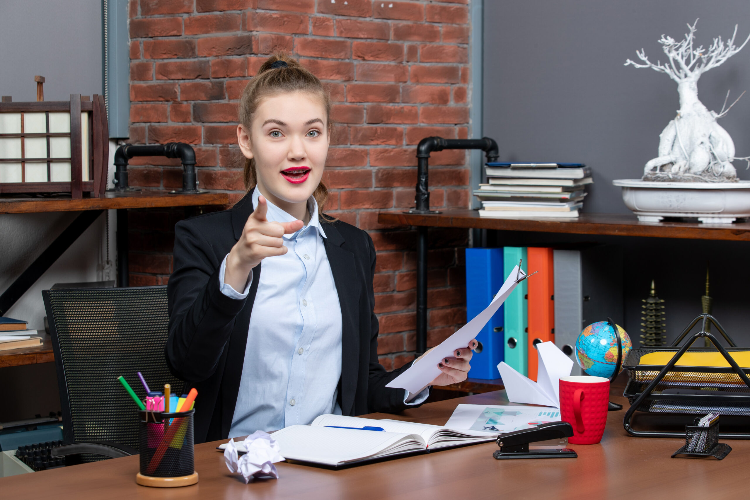 satisfied-positive-young-lady-sitting-table-holding-document-pointing-forward-office